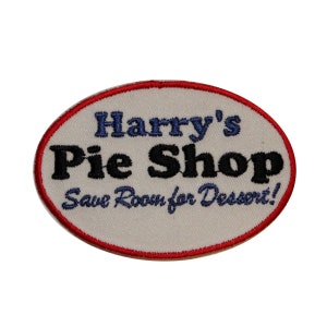 Può includere: Toppa ovale bianca ricamata con cuciture rosse attorno ai bordi. Il testo "Harry's Pie Shop Save Room for Dessert!" è ricamato in blu e nero.