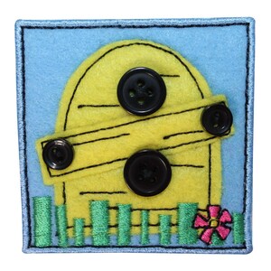 May include: A yellow felt house with a black button door and a green felt grass patch. The house has a black button on the door and a black button on the roof. There is a pink felt flower in the grass.