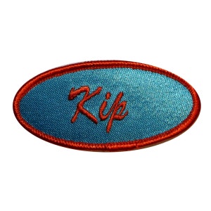 Peut inclure: Un patch en tissu bleu de forme ovale avec une bordure brodée rouge. Le mot "Kip" est brodé en rouge sur le patch.