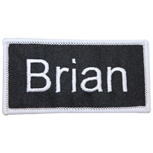 Peut inclure: Écusson rectangulaire en tissu noir avec des coutures blanches autour des bords. Le nom "Brian" est brodé en blanc sur l'écusson.