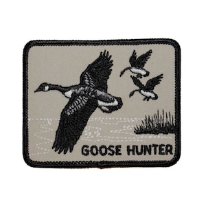 May include: A beige embroidered patch featuring a black goose in flight at the centre. The patch has a black border and the text "GOOSE HUNTER" in black lettering at the bottom.
