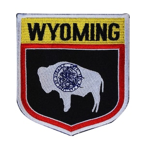 May include: A black and white embroidered patch with a red and yellow border. The patch features a white silhouette of a buffalo with a blue circle containing the state seal of Wyoming. The text "WYOMING" is in black letters on a yellow background.
