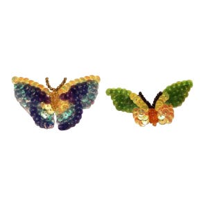 Può includere: Due decorazioni a forma di farfalla colorate fatte di paillettes. Una farfalla è viola, blu e gialla, e l'altra è verde e gialla.