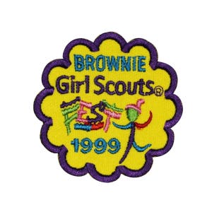 Può includere: Una toppa ricamata gialla con cuciture viola a forma di fiore. La toppa dice "Brownie Girl Scouts® Fest 1999" e presenta un personaggio dei cartoni animati colorato.