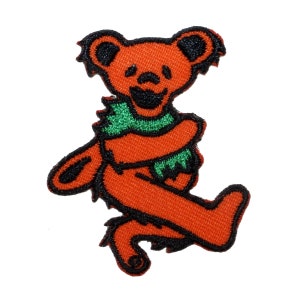 May include: Orange and green embroidered patch of a smiling bear with a green scarf. The bear is standing on its hind legs with its arms crossed.
