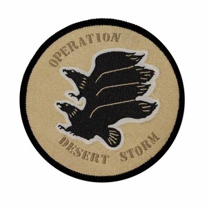 Può includere: Un cerotto rotondo color beige con cuciture nere attorno al bordo. Il cerotto presenta una silhouette nera di un uccello con le ali spiegate. Il testo "OPERATION DESERT STORM" è scritto in beige attorno all'uccello.