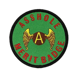 May include: A green and black round embroidered patch with the text "Asshole Merit Badge" in red lettering around the edge. In the center is a brown circle with a gold "A" inside, with gold wings on either side.