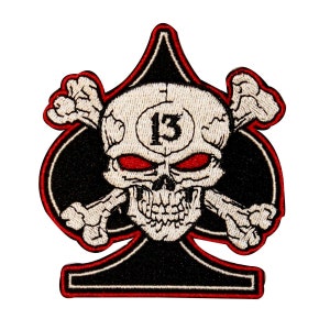 May include: Black and white embroidered patch with a skull and crossbones. The skull has red eyes and a white circle with the number "13" in the center of its forehead. The skull and crossbones are on top of a black spade shape with a red outline.