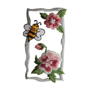 Può includere: Toppa ricamata da stirare con un'ape e dei fiori in una cornice. L'ape è gialla e nera con un viso sorridente. I fiori sono rosa e bianchi con foglie verdi.