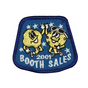May include: Blue embroidered patch featuring two cartoon characters, one wearing sunglasses, giving a high five. The text "2001 BOOTH SALES" is below the characters.