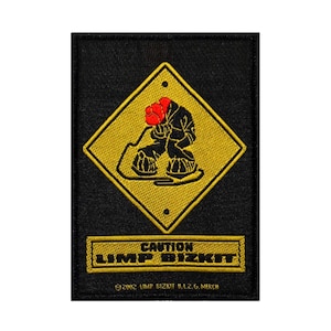 May include: A black and yellow caution sign with a cartoon figure wearing a red hat and black clothing. The text "CAUTION LIMP BIZKIT" is below the sign. The text "©2002 LIMP BIZKIT V.L.L.G. MERCH" is below the text.