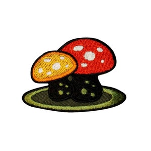 May include: An embroidered patch featuring two mushrooms, one red with white spots and one yellow with white spots, on a green background.