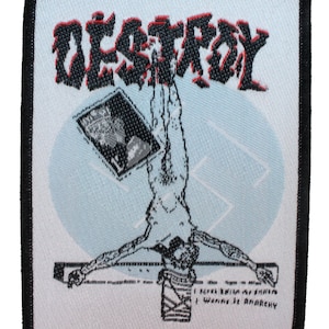 Peut inclure: Écusson brodé avec le mot "DESTROY" en noir et rouge, au-dessus d'une figure inversée sur une croix. L'écusson présente un cercle bleu clair et le texte "I Never Knew Anarchy, I Wanna Be Anarchy."