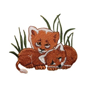 May include: Two brown lion cubs with white faces are cuddling in a patch of green grass. The cubs are embroidered with brown and white thread.