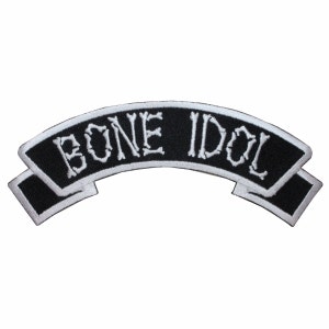 Könnte beinhalten: Schwarz-weiß gestickter Aufnäher mit dem Text "BONE IDOL" in weißer, knochenförmiger Schrift auf einem schwarzen Banner.