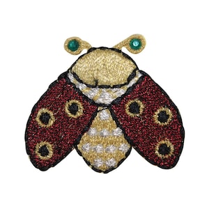 Peut inclure: Un patch brodé en forme de coccinelle rouge et or avec des accents de gemmes vertes. La coccinelle a un corps à motifs blancs et dorés et des points noirs sur ses ailes.