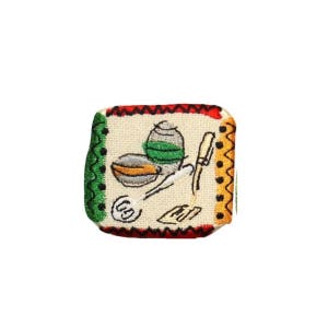 May include: A small, square, embroidered patch with a white background and a red, green, and black border. The patch features a stylized drawing of kitchen utensils, including a bowl, a whisk, a knife, and a spatula.