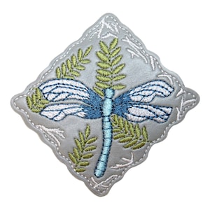 May include: A gray felt patch with a blue and white dragonfly embroidered on it. The dragonfly is surrounded by green embroidered leaves.