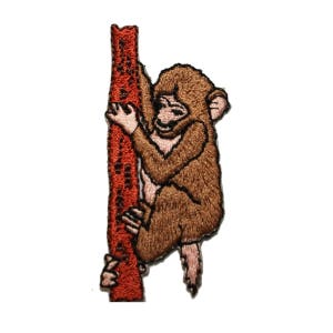 May include: Embroidered patch of a brown monkey climbing a red tree trunk. The monkey has a pink face and is holding onto the trunk with its hands and feet.