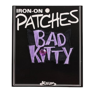 May include: Iron-on patch featuring the text "Bad Kitty" in purple embroidery on a black background. The patch has a purple rhinestone in the centre.