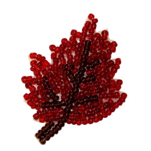 May include: A decorative maple leaf made of red and dark red beads. The beads are arranged on a white backing, creating a textured, three-dimensional effect. The leaf shape is clearly defined, with detailed edges.