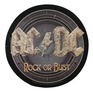 May include: A round black and gray speaker with the words "AC/DC" and "Rock or Bust" in gold lettering. The speaker has a textured surface and a distressed look.