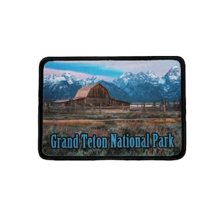 May include: A brown wooden barn with a mountain range in the background. The mountains are covered in snow and the sky is blue. The text "Grand Teton National Park" is printed below the image.