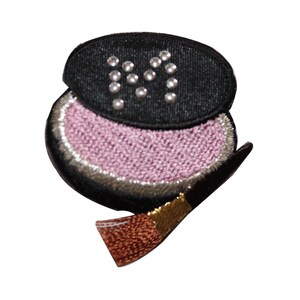 May include: A black and pink embroidered patch of a compact with a brush. The compact has a black lid with white pearl-like embellishments. The compact is open and the inside is pink.