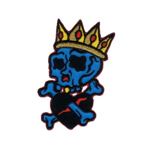 May include: Blue skull wearing a gold crown holding a black heart with crossed bones. The skull has a red mouth and black eyes. The heart has a red outline.