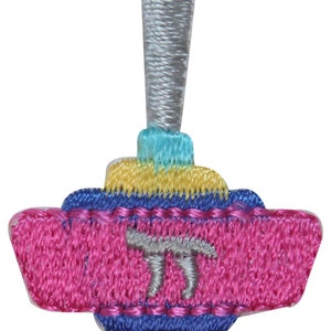 Può includere: Un dreidel ricamato di colore rosa e blu con una cima argentata e la lettera ebraica 'nun' al centro.