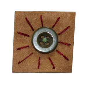 May include: A brown square piece of fabric with a silver button sewn in the centre. The button has a green four-leaf clover design and is surrounded by red stitching.