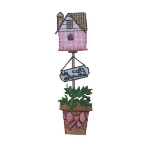 May include: A pink and white birdhouse with a brown roof sits on a wooden post with a sign that reads "No cats allowed". The post is in a brown pot with a pink bow and green plants.