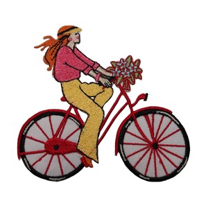Peut inclure: Un patch brodé représentant une femme à vélo rouge avec un bouquet de fleurs dans le panier. La femme porte une chemise rose, un pantalon jaune et un chapeau.