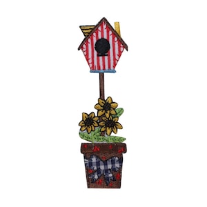 May include: Red and white striped birdhouse with a yellow roof, sitting on a brown post, with a brown pot with a blue and white checked bow, and three yellow sunflowers.