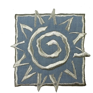 May include: A square, blue fabric patch with a silver embroidered sun design. The sun features a spiral center and radiating lines, with a frayed, silver border. The patch is likely for decorative purposes, such as on clothing or bags.