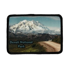 May include: A black and white embroidered patch with a scenic image of a snow-capped mountain range and a dirt road winding through the landscape. The text "Denali National Park" is printed in blue on the patch.