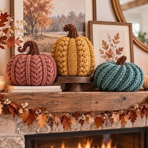 May include: Three handmade crochet pumpkins in muted autumn colors. The left pumpkin is dusty rose with a dark brown stem, the center pumpkin is mustard yellow with a dark brown stem, and the right pumpkin is teal with a dark brown stem.