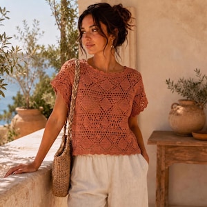 May include: A short-sleeved, terracotta-colored crochet top featuring a geometric diamond pattern throughout the knit. The garment has a round neckline and a scalloped hemline. A woven straw-style shoulder bag with a braided strap is also visible.