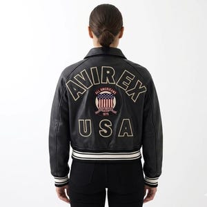 May include: A black leather varsity jacket featuring the text AVIREX in an arched font and USA in a straight font on the back. A circular patch in the center displays the text ALL AMERICANS ESTABLISHED 1975 with a graphic of the American flag.