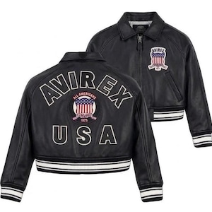 May include: A black leather jacket featuring a front zipper, a collar, and striped ribbed cuffs and hem. The back displays the text AVIREX USA and a circular patch with an American flag and the text ALL AMERICANS ESTABLISHED 1975.
