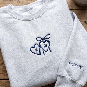 May include: A light gray heather crewneck sweatshirt featuring dark blue embroidery on the chest of two connected hearts with a bow, containing the letters A and B. The right sleeve is embroidered with the date 9-13-26 in dark blue thread.