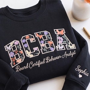 May include: A black crewneck sweatshirt featuring the letters BCBA filled with floral and butterfly embroidery. Below the letters, the text reads Board Certified Behavior Analyst. The sleeve is embroidered with the name Sophia and a small heart.