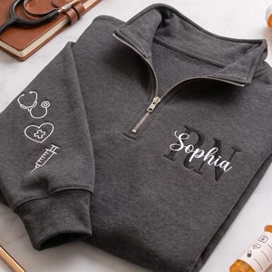 May include: A dark grey quarter-zip sweatshirt featuring the embroidered name Sophia over the letters RN on the chest. The sleeve displays white line art icons of a stethoscope, a heart with a cross, and a syringe. The fabric has a heathered texture.