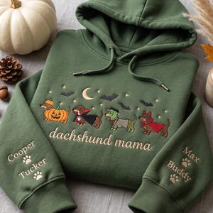 May include: An olive green hooded sweatshirt featuring embroidered Halloween-themed dachshunds, a crescent moon, bats, and stars. The text "dachshund mama" is centered, with the names "Cooper," "Tucker," "Max," and "Buddy" on the sleeves.