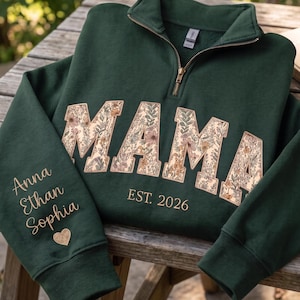 May include: A dark green quarter-zip sweatshirt featuring the word MAMA in a floral print across the chest. Below the text is EST. 2026. The sleeve displays the names Anna, Ethan, and Sophia in gold lettering, followed by a small gold heart icon.