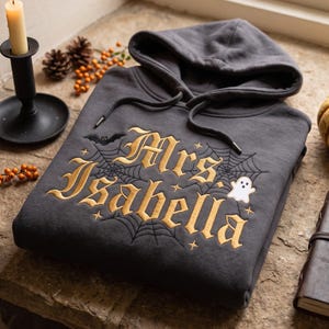 May include: A dark gray hooded sweatshirt featuring gold embroidered text that reads Mrs. Isabella. The design includes a spider web, a small black bat, a white ghost, and several small star accents stitched onto the front of the garment.