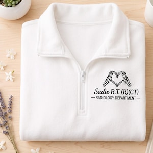 May include: A white quarter-zip pullover featuring a black graphic of two skeleton hands forming a heart shape above the text Sadie R.T. (R)(CT) RADIOLOGY DEPARTMENT. The garment has a high collar with a central zipper closure.