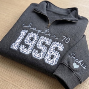 May include: A charcoal gray quarter-zip sweatshirt featuring the embroidered text "chapter 70" and the year "1956" in a floral-patterned applique. The sleeve is embroidered with the name "Sophia" and a small heart icon near the cuff.