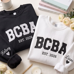 May include: A black sweatshirt with white embroidered text reading BCBA EST. 2026 and a white sweatshirt with black embroidered text reading BCBA EST. 2026. Both feature personalised names and heart icons embroidered on the sleeve cuffs.