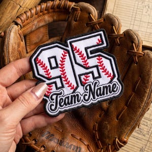 May include: A white embroidered patch featuring the number 95 in a bold font with red baseball-style stitching patterns throughout the digits. Below the number, the words Team Name are stitched in black cursive script with a black border.